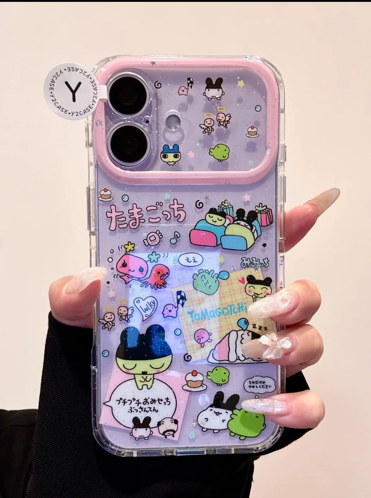 A hand holding a clear phone case with a pink border, featuring Tamagotchi characters, glitter, and stickers on a purple background.