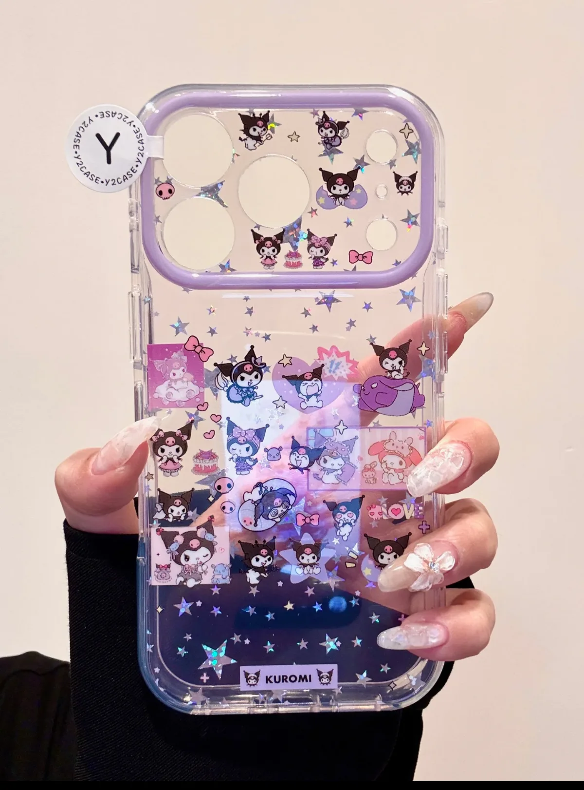 A hand holding a clear phone case with a purple frame, featuring Kuromi characters and glitter stars.