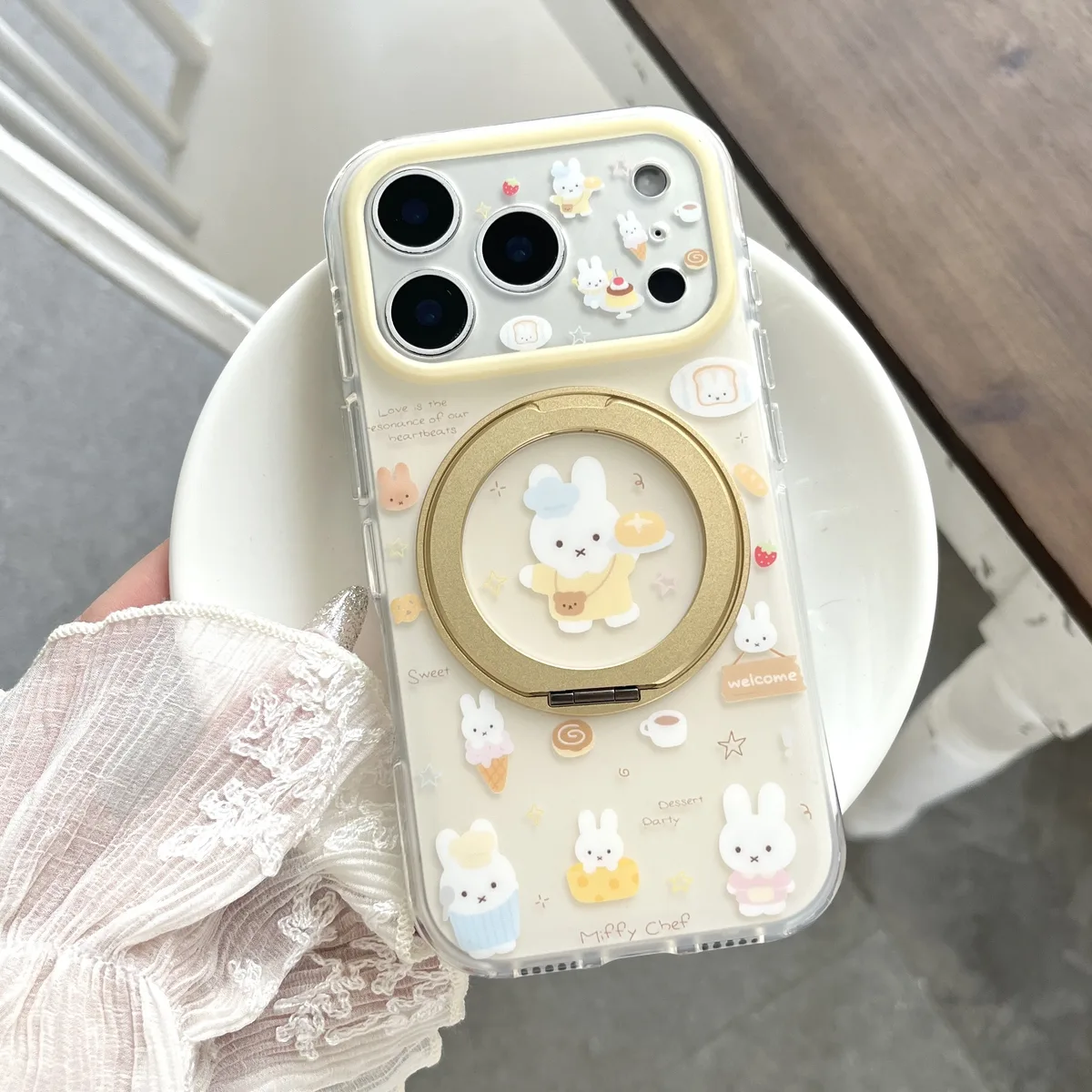 A clear iPhone case with a Miffy chef design and a gold 360° foldable ring stand, shown with a beaded strap.