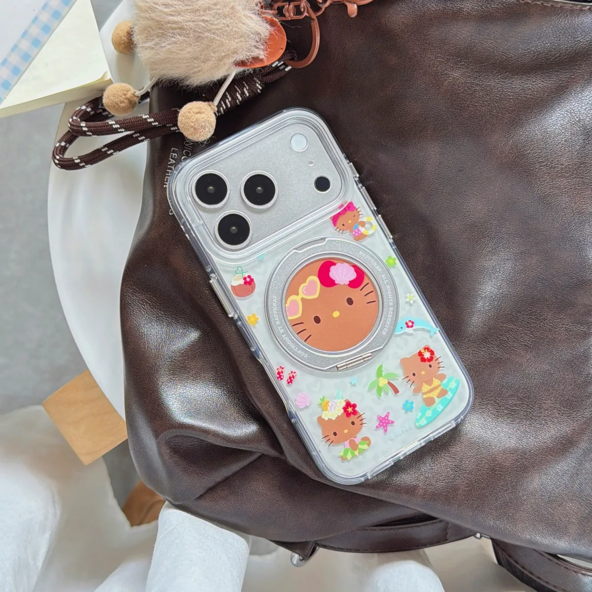 Clear phone case with Hello Kitty beach design and 360 stand, featuring a magnetic ring and a pearl and star charm strap, held in a hand.