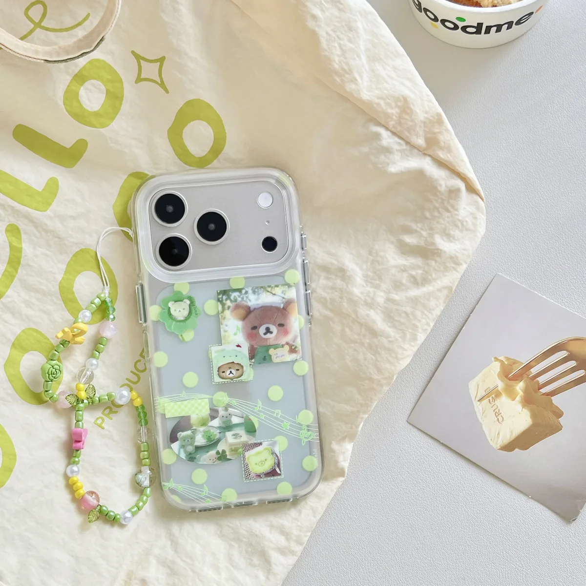 A clear phone case with green polka dots, bear stickers, and music notes, shown with a beaded strap on a white surface.