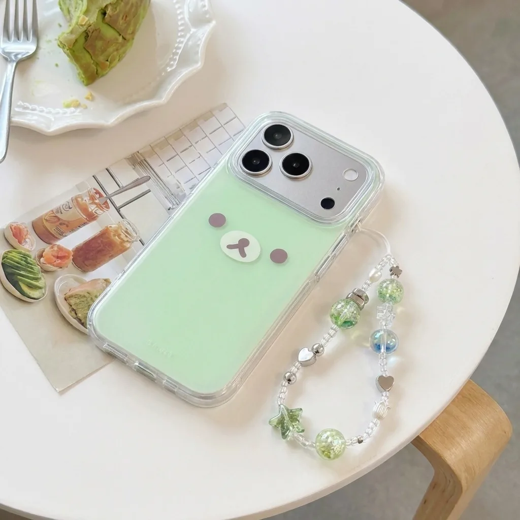 A mint green phone case with a cute bear face design, shown on a white table with a beaded strap and a plate of avocado toast.
