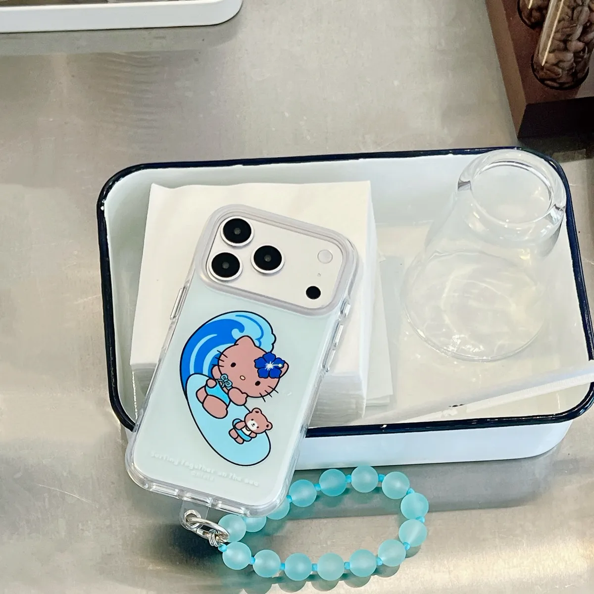 Clear phone case with a blue wave design featuring Hello Kitty surfing, attached to a light blue beaded strap, placed on a white tray.