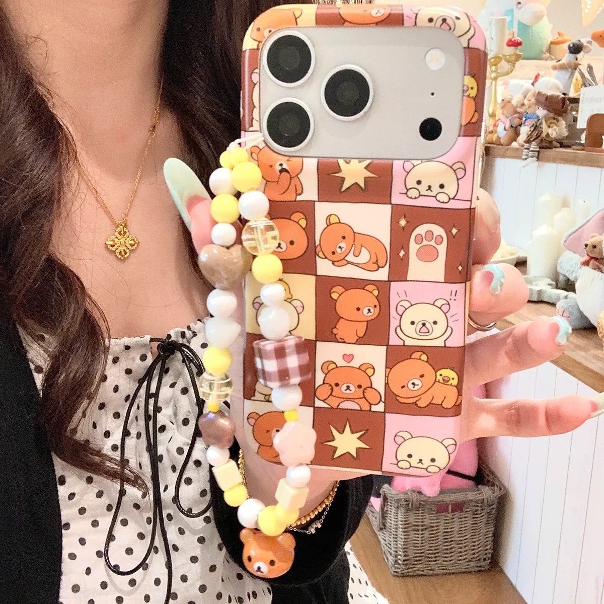 A person holding a phone with a Rilakkuma patchwork case and a beaded strap, showing the back of the phone with camera cutout.