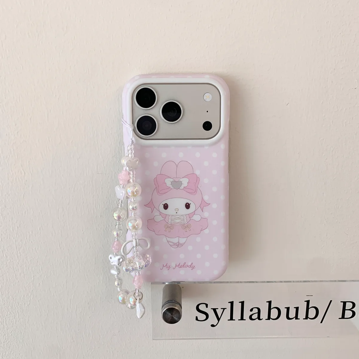 Pink iPhone case with My Melody ballet design and a beaded strap, displayed on a wall and held in hand.