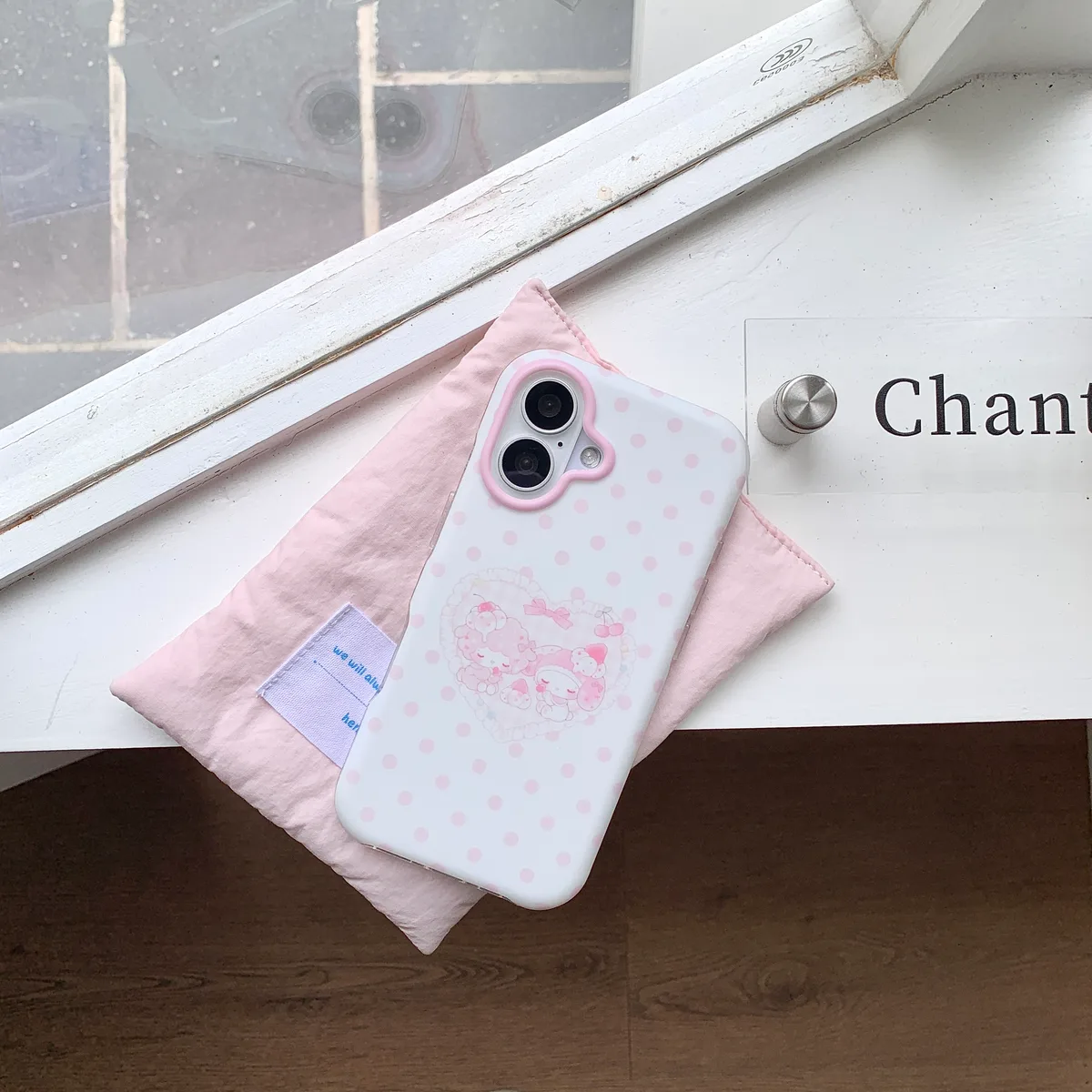 White iPhone case with pink polka dots and My Melody characters, placed on a pink pouch