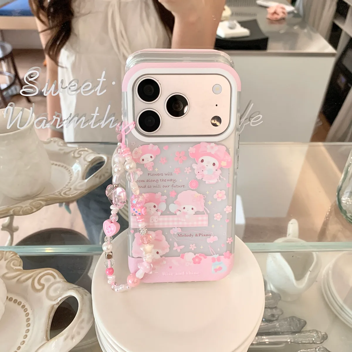 A clear iPhone case with pink border and Melody & Piano design, featuring a beaded strap with hearts and pearls, held