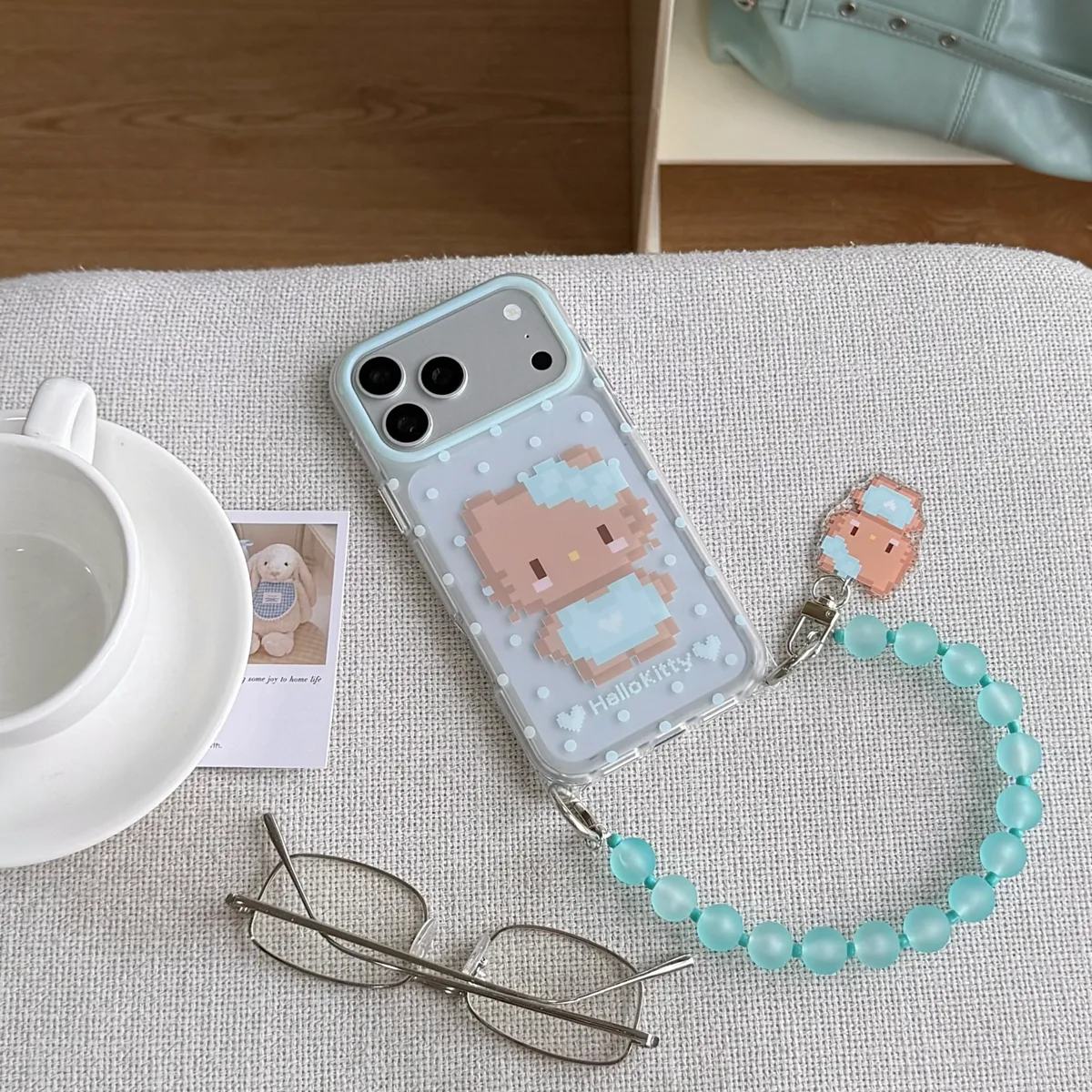 A clear phone case with a pixelated Hello Kitty design and a mint green beaded strap, placed on a white plate and held by a hand.