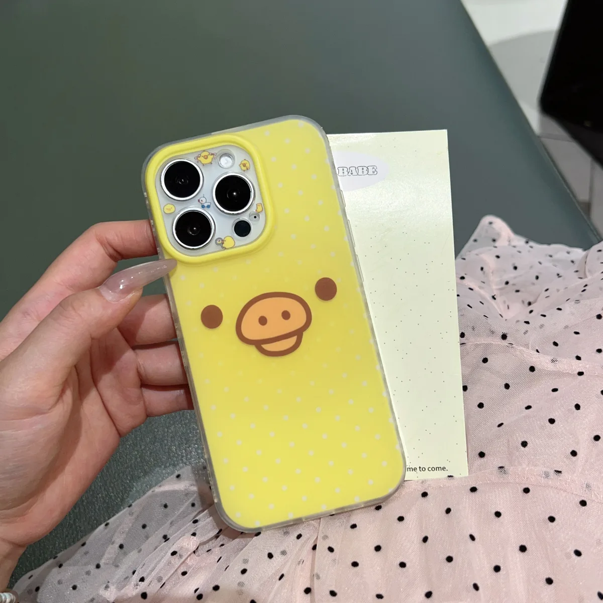 A hand holding a yellow polka dot phone case with a pig face design, featuring a beaded strap and a clear protective edge.