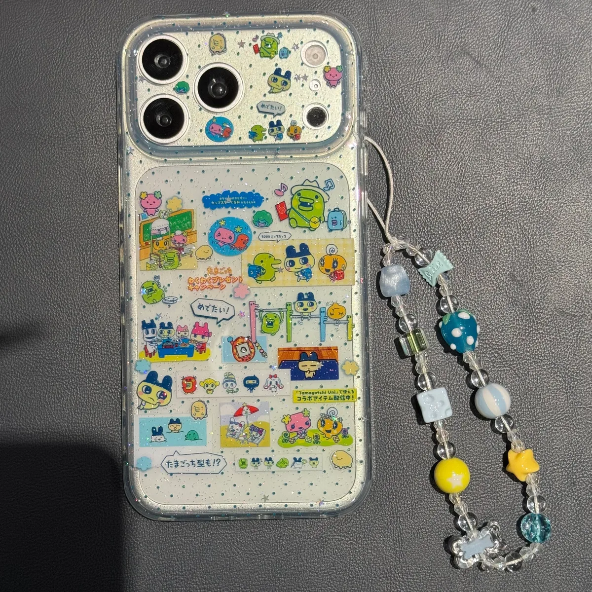 Clear iPhone case with Tamagotchi characters and glitter, featuring a beaded strap, shown on a table and held in hand