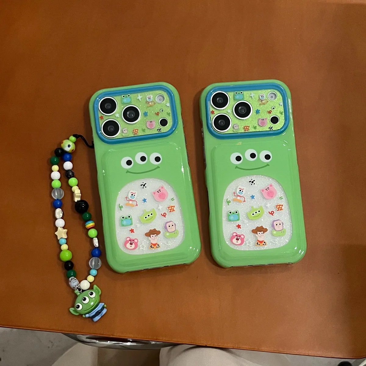 Green phone case with three-eyed alien design and glitter-filled bubble pocket, featuring Toy Story characters and a beaded strap with an alien charm.