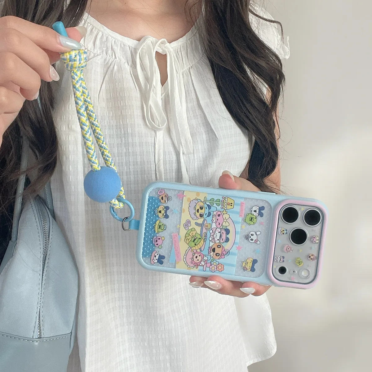 Pastel Tamagotchi phone case with character artwork and a braided blue and yellow wrist strap